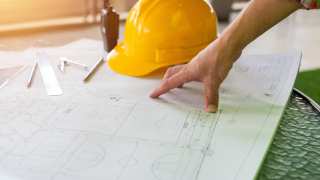 A contractor looking at blueprints with a yellow hardhat on the table.